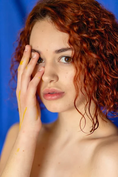 红头发卷曲的女人手涂着黄色的油漆 看着蓝色背景上的相机 艺术或妇女博客的概念摄影 — 图库照片