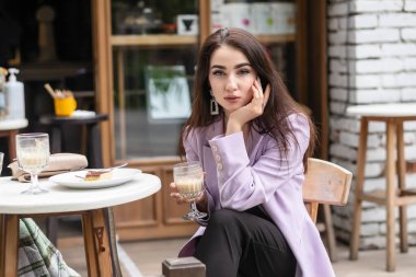 Romantik bir ruh hali içinde, mutlu, pembe ceketli, şık elbiseli, kafede erkek arkadaşını bekleyen, cappuccino içen, mutlu bir kadın.