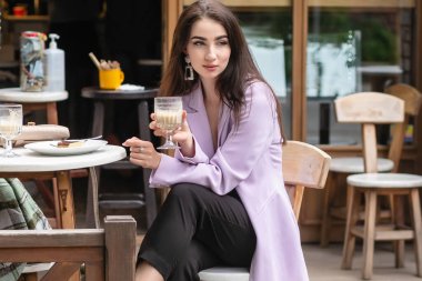 Romantik bir ruh hali içinde, mutlu, pembe ceketli, şık elbiseli, kafede erkek arkadaşını bekleyen, cappuccino içen, mutlu bir kadın.