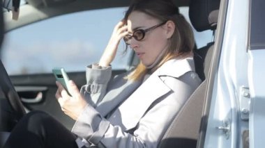Ulaşım, araç ve teknoloji konsepti. Güzel bir genç kadın arabasında otururken akıllı telefon kullanır. Cep telefonuyla iletişim. Sosyal ağlarda sörf