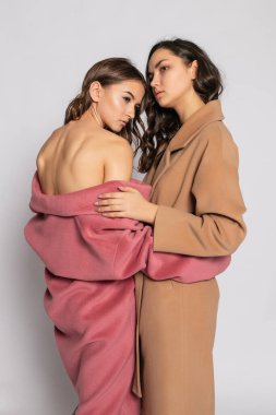 İki gülümseyen esmer kadın modelin moda portresi. Yazın gri arka planda poz veren sıradan hippi paltosu. Tam uzunluk.