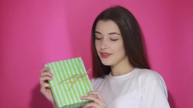 Çekici bir kadın hediyesini yakalıyor ve kutusunu kameraya veriyor, Noel hediyesini paylaşıyor arkadaşça gülümsüyor, tatilleri kutluyor, doğum gününü kutluyor. Stüdyo çekimi pembe arkaplanda izole edildi