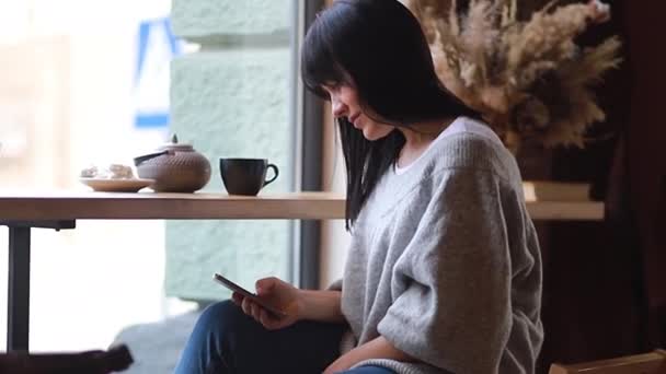 女性はコーヒーを飲み カフェで携帯電話を使う 女の子は一人で居心地の良いコーヒーショップに座って 若い女性はスマートフォンで読んで — ストック動画