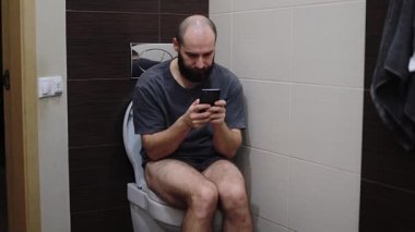 Genç bir adam tuvaletteki tuvalette oturuyor ve akıllı telefona bakıyor. Bir şey onu şaşırttı.