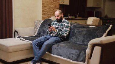 Neşeli orta yaşlı bir adamın modern apartmandaki telefon tebessümünü kullanarak koltukta oturması mesajlaşarak kayıt teknolojisi izole edilmiş yaşam tarzı.
