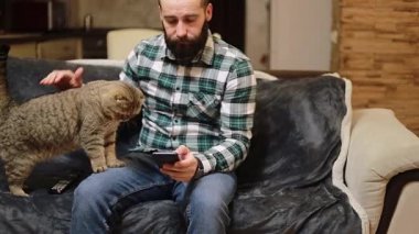 Genç adam akıllı telefon kullanıyor ve rahat bir evde evcil bir kediyle kanepede yatıyor. Süveter giyen gülümseyen bir erkek cep telefonuyla internette sosyal medyada gezinirken oturma odasındaki rahat kanepede dinleniyor..