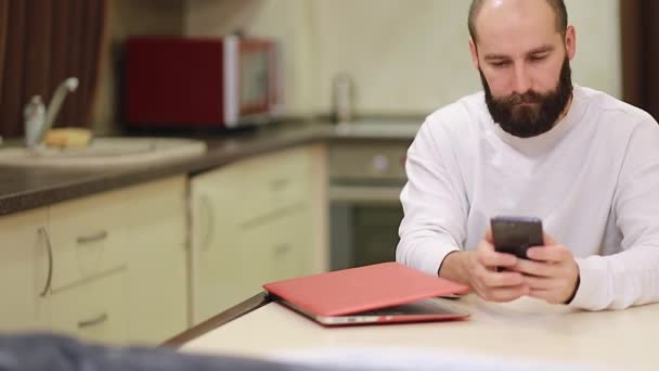 便利な千年独身男は キッチンでテーブルに座っているスマートフォンは ウェブ上でチャットを楽しむ自由な時間を過ごす日付サービス 自宅から電子商取引のウェブサイトで購入を行います 現代のテクノロジーの使い過ぎの概念 — ストック動画
