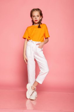 Pembe stüdyo arka planında uzun boylu küçük bir kız. Turuncu tişört, beyaz pantolon ve beyaz spor ayakkabı giyiyor. Şık çocuklar, mutlu çocukluk, duygular. Boşluğu kopyala