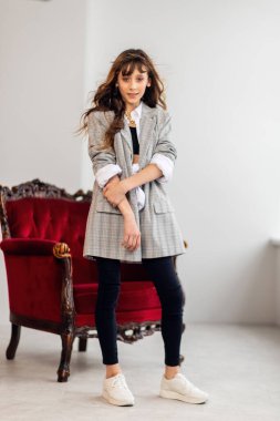 Mutlu küçük kız portresi. Gri ceketli küçük bir kız, çarpık bir iç mekanda, bordo bir koltukla. Beyaz spor ayakkabılar. Çocuk modası, mutlu çocuklar. kız model