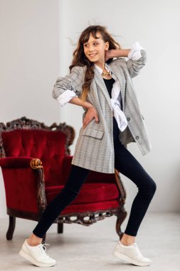 Mutlu küçük kız portresi. Gri ceketli küçük bir kız, çarpık bir iç mekanda, bordo bir koltukla. Beyaz spor ayakkabılar. Çocuk modası, mutlu çocuklar. kız model