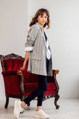 Mutlu küçük kız portresi. Gri ceketli küçük bir kız, çarpık bir iç mekanda, bordo bir koltukla. Beyaz spor ayakkabılar. Çocuk modası, mutlu çocuklar. kız model