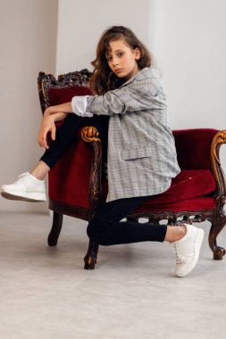 Mutlu küçük kız portresi. Gri ceketli küçük bir kız, çarpık bir iç mekanda, bordo bir koltukla. Beyaz spor ayakkabılar. Çocuk modası, mutlu çocuklar. kız model