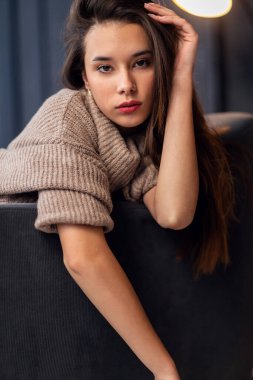 Güzel bir genç kız bir sandalyenin arkasında oturuyor. Kız kahverengi, rahat bir kazak giyiyor. Eli onun kalın, dalgalı saçlarını düzleştiriyor..