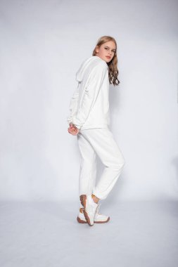 Gri arka planda izole edilmiş beyaz eşofmanlı güzel sarışın kadın. Beyaz eşofmanlı sarışın kız beyaz bir arka planda duruyor.