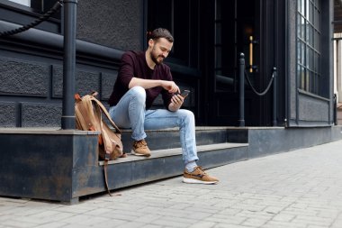 Sakallı, modern aletlerle gezen mutlu genç adam. basamaklarda oturuyor ve bir cep telefonuna bakıyor.