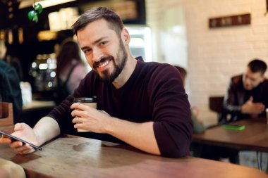 Genç hippi barda cep telefonuyla mesajlaşıyor ve cappuccino içiyor. Genç moda adamı / hipster öğle yemeği saatinde kafede espresso içiyor ve tablet bilgisayar üzerinde çalışıyor.
