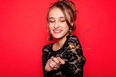 Havai fişekler ve insan konsepti - gülümseyen genç kadın ya da genç kız maytaplı mutlu kadın kırmızı arka planda siyah elbiseyle kutluyor. Kız ateşe bakıyor, şaşırmış.