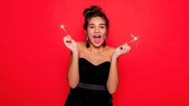 Havai fişekler ve insan konsepti - gülümseyen genç kadın ya da genç kız maytaplı mutlu kadın kırmızı arka planda siyah elbiseyle kutluyor. Kız kameraya gülümsüyor, seviniyor.