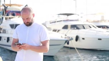 Handsome Caucasian man smiles while using cell phone as he leans against railing at end of breakwater of bay near boats and yachts parking