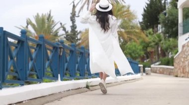 Gülümseyen, güzel bir genç kadın, güneş gözlüklü, açık yaz elbisesi, rüzgarda dalgalanan bir rüzgar, ve güneş şapkası, sokak boyunca yürüyen, palmiye ağaçları, deniz kenarında. Sıcak yaz günü, Ağır çekim