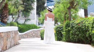 Beyaz elbiseli genç bir kadın elinde şapkasıyla Yunan sokaklarında yürüyor.