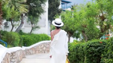 Beyaz elbiseli genç bir kadın elinde bir şapkayla Yunan İHA 'sının caddelerinde yürüyor.