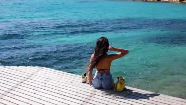慢动作 年轻女子坐在木制码头上 双腿悬在碧绿的水面上 — 图库视频影像