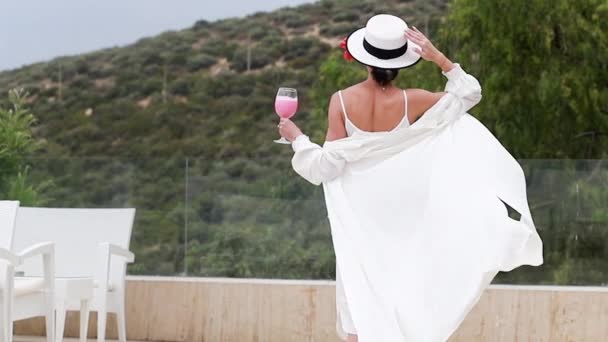 身穿白衣 头戴礼帽的女孩带着鸡尾酒在度假胜地的阳台上走着 奢华旅行或高档生活方式 — 图库视频影像