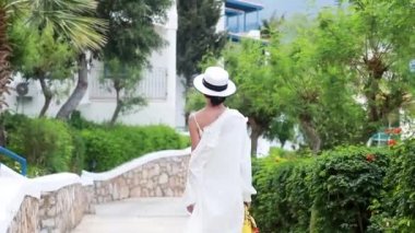 Beyaz elbiseli genç bir kadın elinde bir şapkayla Yunan İHA 'sının caddelerinde yürüyor.