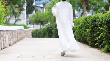 Beyaz elbiseli genç bir kadın elinde şapkasıyla Yunan sokaklarında yürüyor.