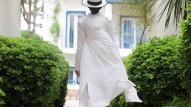 Beyaz elbiseli, elinde hasır şapkası olan genç bir kadın oteli gözetliyor..