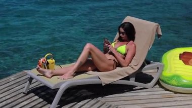 Turkuaz denizde cep telefonu kullanan şamandıradaki tatilci kadın. Kız, bikinili tatil köyünde dinleniyor ve tatilin tadını çıkarıyor. Sosyal medyada sohbet.