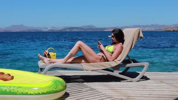 5G-Telefon schnelle Internetverbindung auf Urlauberin auf Liege schwimmt mit Handy in türkisfarbenem Meer. Mädchen entspannen sich und genießen den Urlaub im Resort im Bikini.