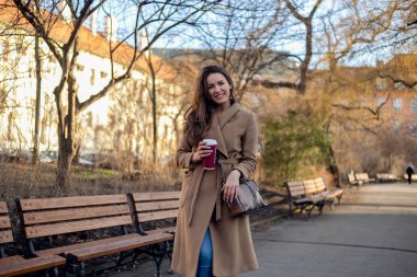 Sıcak kahveyle bankta dinlenen zarif bir kadın, sonbahar şehri arka planı, sıcak melodiler ve rahat bir ruh hali..