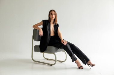 Modern sandalyede oturan, beyaz arka planda, asgari düzeyde siyah elbiseli zarif bir kadının tam vücut portresi..