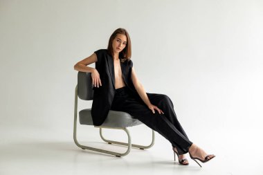 Minimum stüdyo portresi, yan koltukta oturan siyahlı kadın, beyaz zeminde kendine güvenen sakin ruh hali..