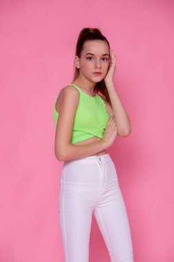 Düşünceli duruşu olan yarım vücut portresi, neon yeşil üst ve pembe arka planda beyaz pantolon..