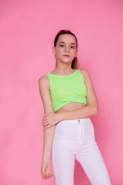 Tam vücut portresi, rahat duruş, neon yeşil üst, beyaz pantolon, pembe arka plan..