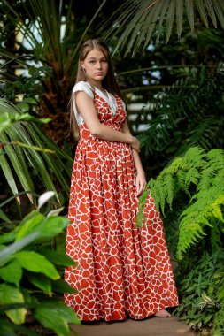 Büyük yaprakların yanında bütün vücut portresi, kırmızı desenli elbise, tropikal sera havası..