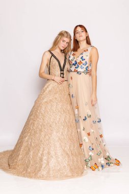 Two women posing together in studio: one in champagne ballgown and one in floral embellished gown, bridesmaid or gala fashion concept.