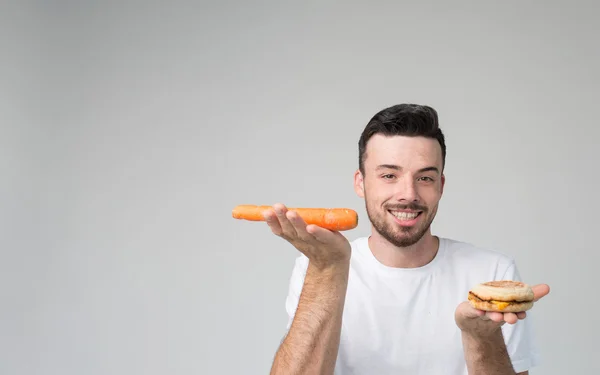Hombre barbudo con una camisa blanca sobre un fondo claro sosteniendo una hamburguesa y una zanahoria — Foto de Stock