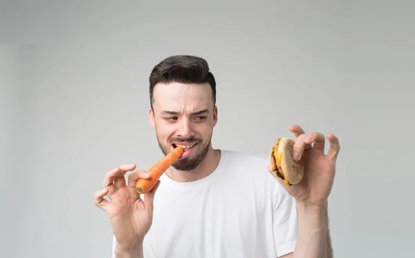 Bebaarde man in een wit overhemd op een lichte achtergrond holding een hamburger en een wortel — Stockfoto