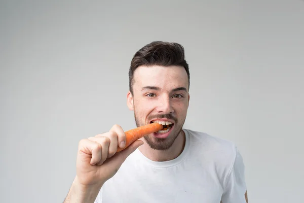 Hombre barbudo con una camisa blanca sobre un fondo claro sosteniendo una hamburguesa y una zanahoria — Foto de Stock