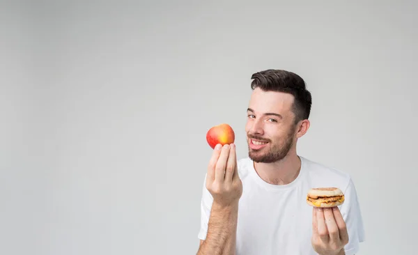Homem barbudo em uma camisa branca em um fundo leve segurando um hambúrguer e uma maçã — Fotografia de Stock