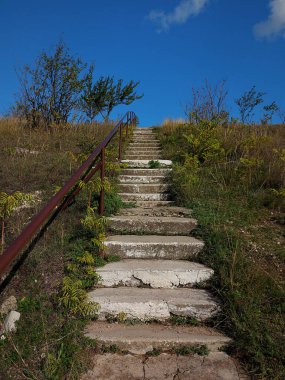 Mavi gökyüzüne çıkan paslı demir boru saplı eski beton merdivenler. Ağaçları ve yeşil otları olan yüksek bir tepe. Güzel bir yer. Kırsal biçim.
