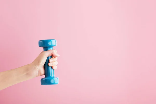 Persons hand lifting blue dumbbell. Fitness equipment for home and gym workout. Train indoors. Sport and healthy lifestyle concept. Copy space in right side. Isolated on pink background.