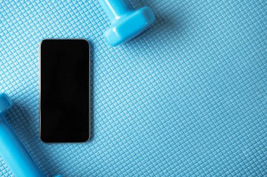 Dambılı spor minderinin üzerinde boş ekranı olan modern akıllı telefonun üst görüntüsü. Cep telefonu ekranında metin için yer. Flatlay tarzı ve kopyalama alanı. Online eğitim. Evde spor yapmak için spor malzemeleri.