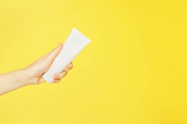 El ve tırnak kremi beyaz plastik bir kapta, kadın elinin sarı arka plan izolasyonunda. Vücut bakım losyonu. Basit boş ambalaj güzellik ürünleri. Model yap, boşluğu kopyala.