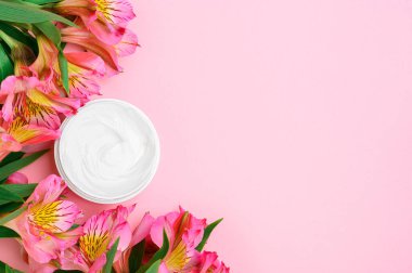 Beyaz bir kavanozda nemlendirici, pembe bir arka planda, alstroemeria çiçekleri ve yeşillikler. Açık plastik ambalajda kremsi bir doku. Besleyici bir kozmetik güzellik ürünüyle kompozisyon.