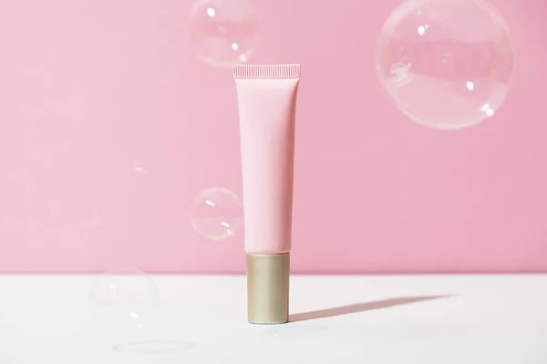 Pink tube with soft eye cream with golden cap on white pink background with soap bubbles. Cosmetic female face care product in plastic packaging. Summer skin cleanser concept. Copy space.